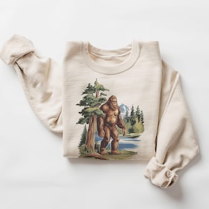May include: A beige sweatshirt featuring a graphic print of Bigfoot walking through a forest. The graphic is in shades of brown, green, and blue.