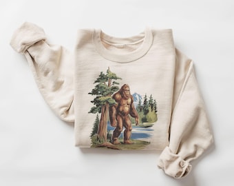 Puede incluir: Una sudadera beige con un estampado gráfico de un Bigfoot caminando por un bosque. El gráfico está en tonos de marrón, verde y azul.