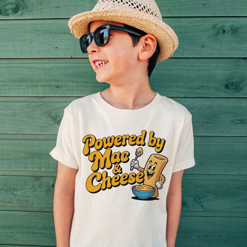 K&ouml;nnte beinhalten: Cremefarbenes T-Shirt mit dem Text "Powered by Mac & Cheese" in Gelb und Orange. Die Grafik zeigt eine l&auml;chelnde Makkaroni-Nudel, die einen L&ouml;ffel &uuml;ber einer Sch&uuml;ssel Makkaroni und K&auml;se h&auml;lt. Das T-Shirt wird von einem Kind getragen.