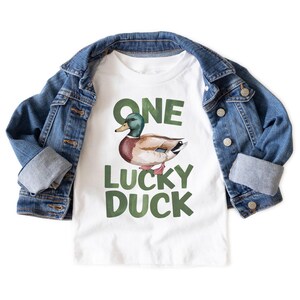 Puede incluir: Camiseta blanca con una ilustraci&oacute;n de pato y el texto "ONE LUCKY DUCK" en verde. Una chaqueta vaquera azul est&aacute; sobre la camiseta. La camiseta es probablemente para un ni&ntilde;o.