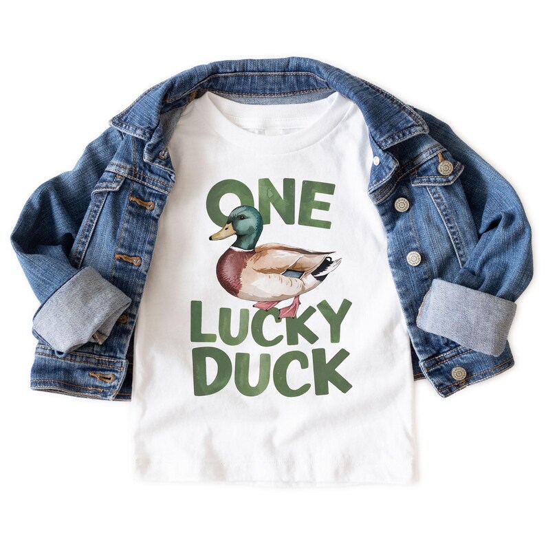 Puede incluir: Camiseta blanca con una ilustraci&oacute;n de pato y el texto "ONE LUCKY DUCK" en verde. Una chaqueta vaquera azul est&aacute; sobre la camiseta. La camiseta es probablemente para un ni&ntilde;o.