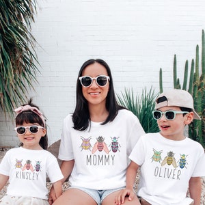 May include: Three white t-shirts with colorful bug illustrations and the names "Mommy", "Kinsley", and "Oliver" printed on them. The shirts are being worn by a woman and two children.