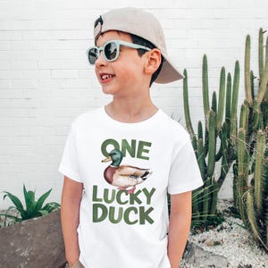 Puede incluir: Camiseta blanca con una ilustraci&oacute;n de pato y el texto "ONE LUCKY DUCK" en verde. La camiseta la lleva una persona con gafas de sol y una gorra de b&eacute;isbol.