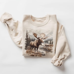 May include: A beige sweatshirt with a brown moose graphic. The moose is standing in a forest setting with trees and a river.