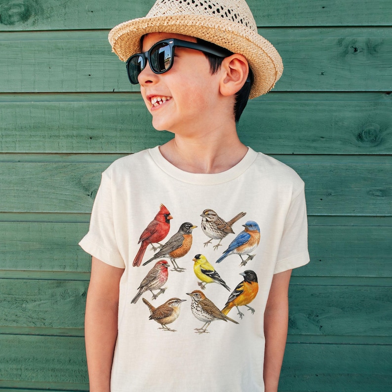 以下が含まれることがあります： さまざまな鳥のカラフルなイラストが描かれたクリーム色のTシャツ。鳥は円形に配置されています。画像内の人物は麦わら帽子とサングラスを着用しています。