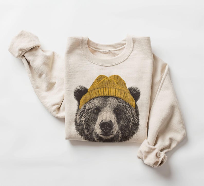 May include: Cream-colored sweatshirt featuring a detailed illustration of a bear's face wearing a mustard yellow beanie. The bear's fur is depicted in shades of brown and black. The sleeves are rolled up, and the sweatshirt is laid flat on a white surface.