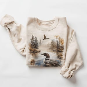 May include: Beige sweatshirt featuring a nature scene with a loon swimming in a lake, trees, and a flying bird. The design is a watercolor painting style. The sweatshirt has long sleeves and a crew neck.