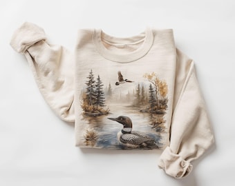 Puede incluir: Sudadera beige con una escena de la naturaleza que representa un somorgujo en un lago, árboles y un pájaro volando. El diseño es de estilo acuarela. La sudadera tiene mangas largas y cuello redondo.