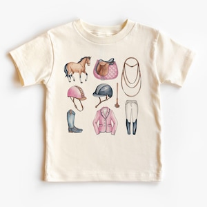 Puede incluir: Una camiseta de color crema con una ilustración de acuarela de equipo de equitación. La ilustración incluye un caballo marrón, una silla de montar rosa, un casco rosa, una fusta, un par de botas de montar y una chaqueta de montar rosa.