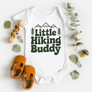 May include: White baby onesie with the text "Little Hiking Buddy" in dark green. The design includes mountain and tree graphics. The image also shows a pair of brown baby shoes and a wooden toy.