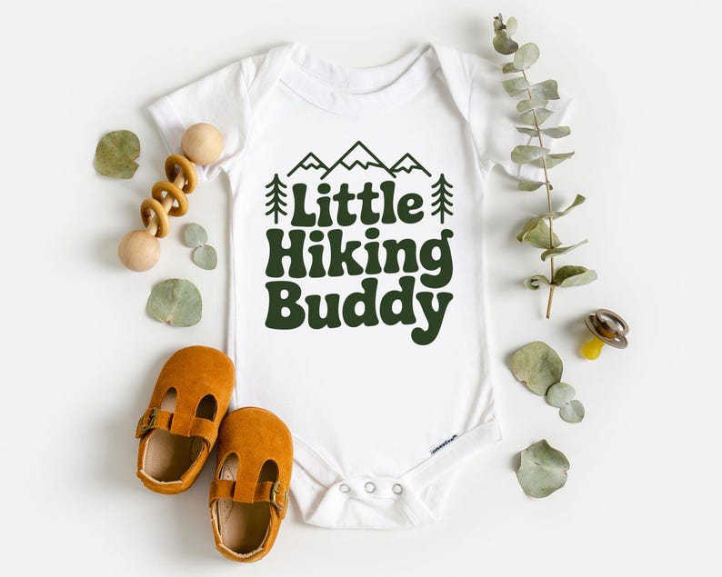 May include: White baby onesie with the text "Little Hiking Buddy" in dark green. The design includes mountain and tree graphics. The image also shows a pair of brown baby shoes and a wooden toy.