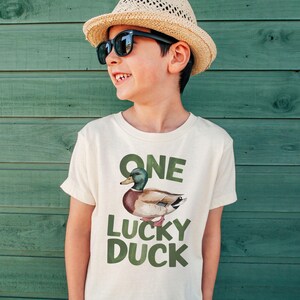 Puede incluir: Una camiseta de color beige claro con el texto "ONE LUCKY DUCK" en letras verdes. La camiseta presenta una ilustraci&oacute;n de acuarela de un pato real. La persona de la imagen lleva un sombrero de paja y gafas de sol.