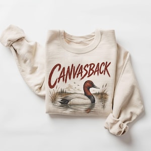May include: A cream-colored crewneck sweatshirt featuring a graphic print of a Canvasback duck swimming in a marsh. The word CANVASBACK is printed in large, dark red, distressed-style lettering above the duck illustration, which includes reeds.