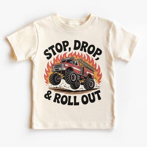 May include: Cream-colored toddler t-shirt featuring a graphic of a red and white monster truck fire engine jumping over flames. The text "STOP, DROP, & ROLL OUT" is printed above and below the truck in black.