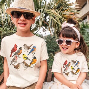 以下が含まれることがあります： クリーム色のTシャツを着た2人の子供たち。カラフルな鳥のイラストが描かれています。赤いカージナル、ルリビタキ、黄色いフィンチなど、さまざまな種類の鳥が描かれています。一人は麦わら帽子とサングラスを、もう一人はサングラスとピンクのヘアバンドを着用しています。