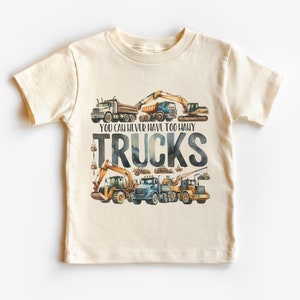 May include: Cream-colored short-sleeved t-shirt featuring a watercolor illustration of various trucks. The text "YOU CAN NEVER HAVE TOO MANY TRUCKS" is printed in a dark blue font. The design includes dump trucks, excavators, and other construction vehicles.