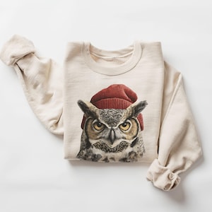 May include: A cream-colored crewneck sweatshirt featuring a detailed graphic print of a great horned owl wearing a dark red knit beanie. The owl has yellow eyes, brown and gray feathers, and a focused expression centered on the chest area.