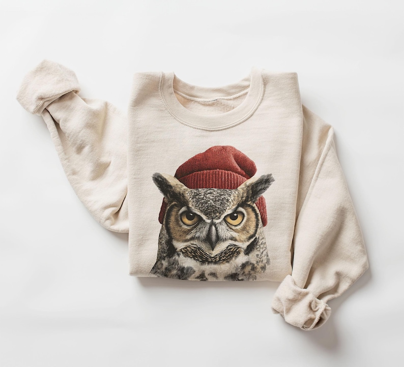 May include: A cream-colored crewneck sweatshirt featuring a detailed graphic print of a great horned owl wearing a dark red knit beanie. The owl has yellow eyes, brown and gray feathers, and a focused expression centered on the chest area.