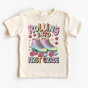 Może przedstawiać: Kremowy t-shirt z kolorową grafiką rolek i napisem "Rolling into First Grade". Projekt zawiera pastelowe kolory, kwiaty i gwiazdki, tworząc zabawną i nostalgiczną atmosferę.