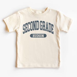 May include: A cream-coloured short-sleeved t-shirt featuring the navy blue text "SECOND GRADE" arched across the top, with the name "HUDSON" in a navy blue oval below. The shirt is made of a soft material.