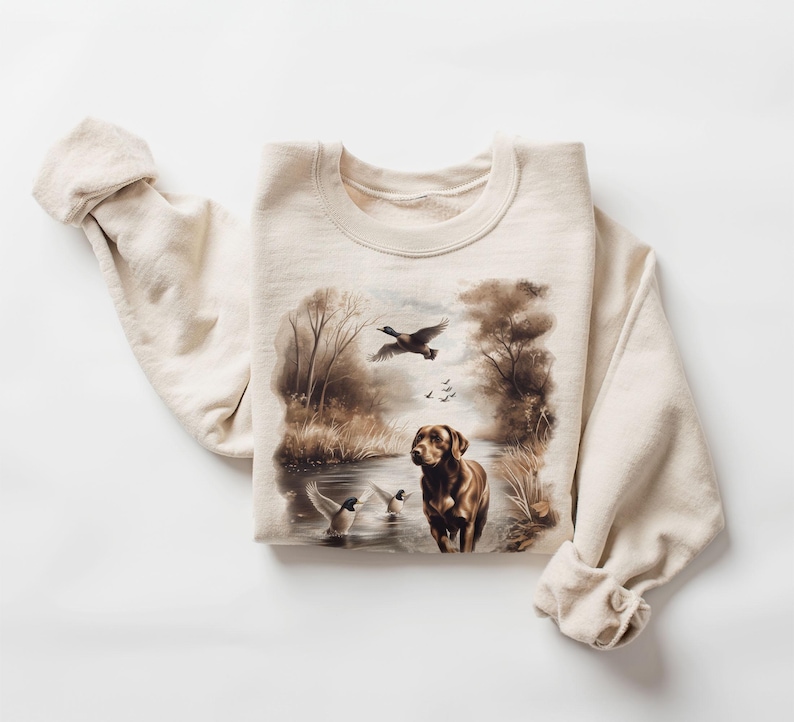 Puede incluir: Una sudadera beige con una ilustraci&oacute;n marr&oacute;n y blanca de un perro y patos en un entorno de bosque.