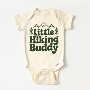 May include: Cream-colored baby onesie with short sleeves. The front features the text "Little Hiking Buddy" in dark green, along with a mountain and tree graphic. The onesie has snap closures at the bottom.