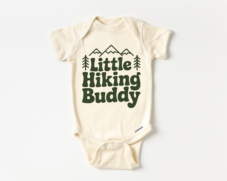 May include: Cream-colored baby onesie with short sleeves. The front features the text "Little Hiking Buddy" in dark green, along with a mountain and tree graphic. The onesie has snap closures at the bottom.