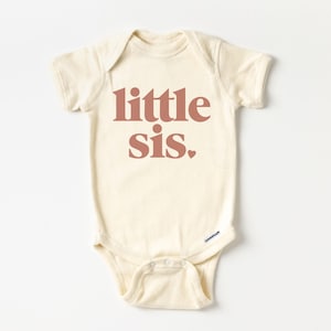 May include: A cream-coloured baby onesie with short sleeves and the text "little sis." printed in a rust-coloured font.
