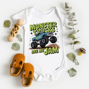 Puede incluir: Body de beb&eacute; blanco con un gr&aacute;fico de un cami&oacute;n monstruo azul y el texto "MONSTER TRUCKS ARE MY JAM" en verde y negro. La imagen incluye un par de zapatos de beb&eacute; naranjas y un juguete de madera.
