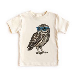 May include: A cream-colored short-sleeve t-shirt featuring a detailed graphic illustration of a brown and white owl wearing bright blue-rimmed glasses. The owl is depicted in a standing position with its head turned toward the viewer.