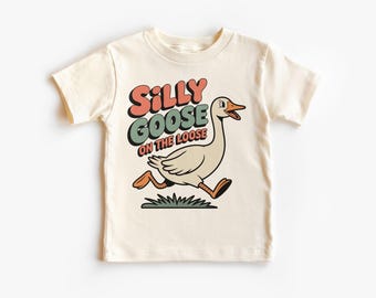 May include: A cream-coloured t-shirt with a cartoon goose running right. The text "Silly Goose on the Loose" is above the goose in a retro font with red, green, and black accents. The goose has orange feet and a yellow beak.