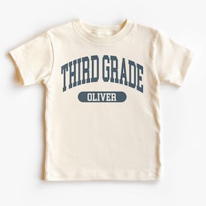 May include: Cream-colored short-sleeved t-shirt with the text "THIRD GRADE" in navy blue arched across the top. Below, a navy blue oval reads "OLIVER". The shirt is made of a soft material, suitable for children.