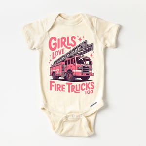 K&ouml;nnte beinhalten: Cremefarbener Baby-Body mit einer rosa Feuerwehrauto-Grafik und dem Text "Girls Love Fire Trucks Too". Das Feuerwehrauto hat eine Leiter und ist vor einem wei&szlig;en Hintergrund platziert. Der Body hat kurze &Auml;rmel.