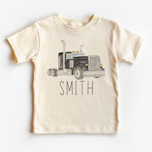 Puede incluir: Una camiseta de color crema con una ilustración de acuarela negra y gris de un camión de plataforma. El texto "SMITH" está impreso debajo del camión.
