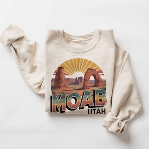 May include: Cream-colored sweatshirt featuring a Moab, Utah graphic. The graphic depicts a desert landscape with arches, a setting sun, and the word "MOAB" in a retro font. The word "UTAH" is below the word "MOAB".