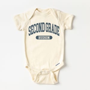 May include: A cream-coloured baby onesie featuring the navy blue text "SECOND GRADE" arched across the top, with "HUDSON" in a navy blue rectangle below. The onesie has short sleeves and snap closures.