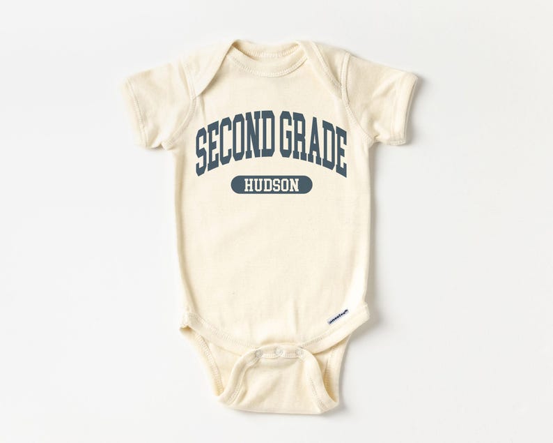 May include: A cream-coloured baby onesie featuring the navy blue text "SECOND GRADE" arched across the top, with "HUDSON" in a navy blue rectangle below. The onesie has short sleeves and snap closures.