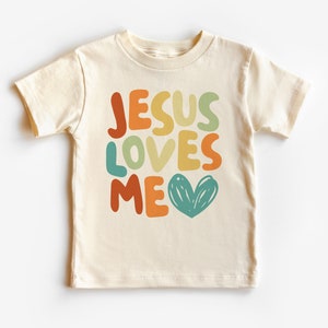 Op de afbeelding: Crèmekleurig T-shirt met korte mouwen met de tekst "JESUS LOVES ME" in kleurrijke, afgeronde letters. Een turquoise hartafbeelding staat onder de tekst. Het shirt is waarschijnlijk gemaakt van zacht katoen.