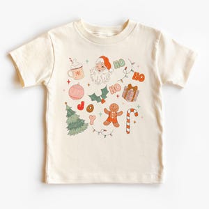 Puede incluir: Camiseta color crema con un dise&ntilde;o navide&ntilde;o. El gr&aacute;fico incluye a Pap&aacute; Noel, un &aacute;rbol de Navidad, un hombre de jengibre, un bast&oacute;n de caramelo y la palabra "HO" repetida. Otros elementos incluyen una taza de chocolate caliente y un regalo envuelto.
