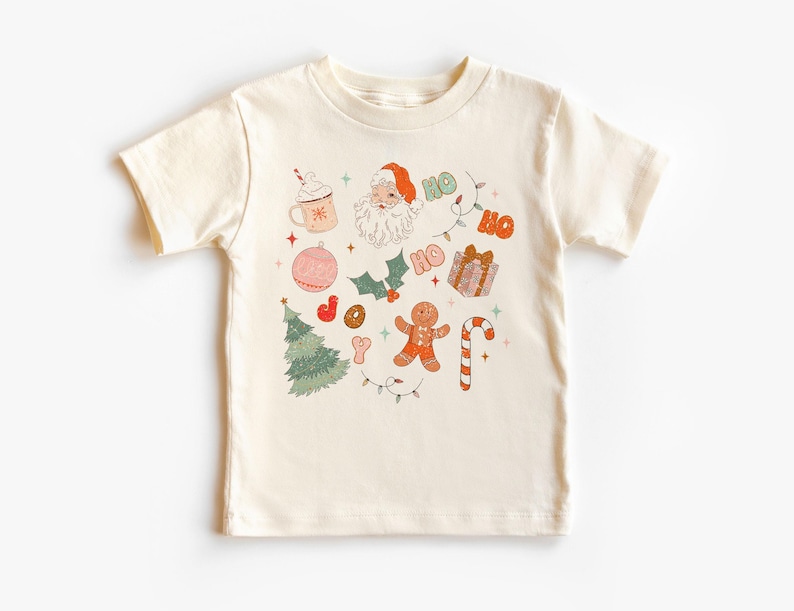 Puede incluir: Camiseta color crema con un dise&ntilde;o navide&ntilde;o. El gr&aacute;fico incluye a Pap&aacute; Noel, un &aacute;rbol de Navidad, un hombre de jengibre, un bast&oacute;n de caramelo y la palabra "HO" repetida. Otros elementos incluyen una taza de chocolate caliente y un regalo envuelto.