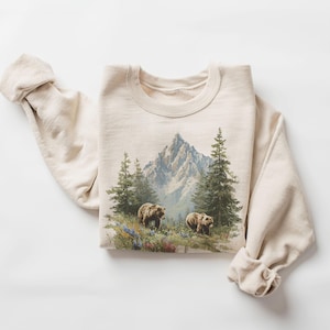May include: Beige sweatshirt featuring a mountain scene with two bears, evergreen trees, and wildflowers. The graphic design is centered on the front of the folded sweatshirt. The sleeves are rolled up. The sweatshirt is made of a soft material.