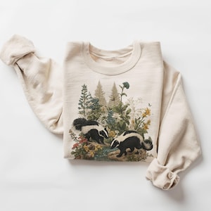 Peut inclure: Sweat-shirt crème avec une scène de nature représentant deux blaireaux et diverses plantes et fleurs. Les blaireaux sont noirs et blancs, et les plantes présentent des teintes vertes, jaunes et bleues. Les manches sont retroussées.