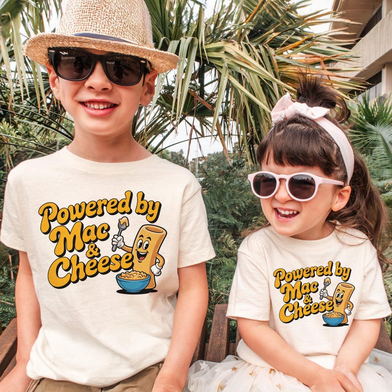 K&ouml;nnte beinhalten: Zwei Kinder tragen cremefarbene T-Shirts mit dem Text "Powered by Mac & Cheese" in Gelb und Orange. Die Shirts zeigen eine Cartoon-Makkaroni-Figur, die einen L&ouml;ffel &uuml;ber einer Sch&uuml;ssel Makkaroni und K&auml;se h&auml;lt. Ein Kind tr&auml;gt einen Strohhut und eine Sonnenbrille.