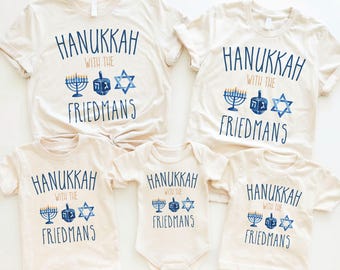 May include: A set of five cream-colored shirts and a baby bodysuit featuring the blue text HANUKKAH WITH THE FRIEDMANS. The design includes a blue menorah, a dreidel with Hebrew letters, and a Star of David in a watercolor style.