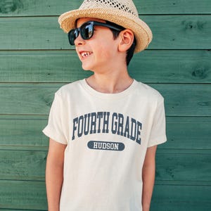 May include: A cream-colored Fourth Grade t-shirt with the name "Hudson" printed below the text. The shirt is worn by a person wearing a straw hat and black sunglasses. The background is a green wooden wall.