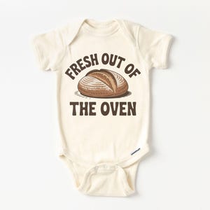 Puede incluir: Body de bebé color crema con el texto "FRESH OUT OF THE OVEN" en letras marrones. Una ilustración marrón de una hogaza de pan está centrada encima del texto. El body tiene mangas cortas y corchetes en la parte inferior.