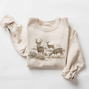 Könnte beinhalten: Ein cremefarbenes Sweatshirt mit Rundhalsausschnitt und einer detaillierten grafischen Illustration von drei Hirschen in einer Naturlandschaft. Das Motiv zeigt einen Hirsch mit Geweih neben zwei weiteren grasenden Tieren.