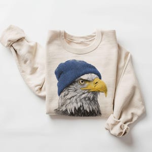 Könnte beinhalten: Cremefarbenes Sweatshirt mit einer detaillierten Illustration eines Adlers mit blauer Mütze. Der Adler hat einen gelben Schnabel und stechende Augen. Die Ärmel sind hochgekrempelt, und das Sweatshirt liegt flach.