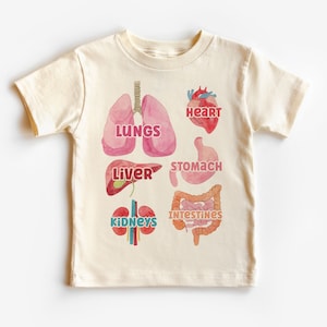 K&ouml;nnte beinhalten: Ein beiges T-Shirt mit einer bunten Illustration der inneren Organe des menschlichen K&ouml;rpers. Die Organe sind mit den Worten "Lungen", "Herz", "Leber", "Magen", "D&auml;rme" und "Nieren" beschriftet.