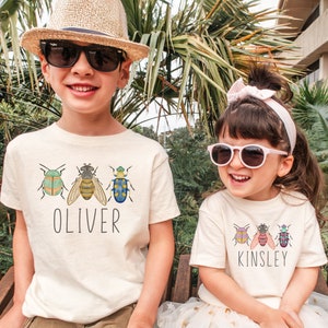 May include: Two children wearing white t-shirts with colorful bug illustrations and their names, "Oliver" and "Kinsley", printed on the front. The boy is wearing a brown hat and sunglasses, while the girl is wearing a pink headband and sunglasses.